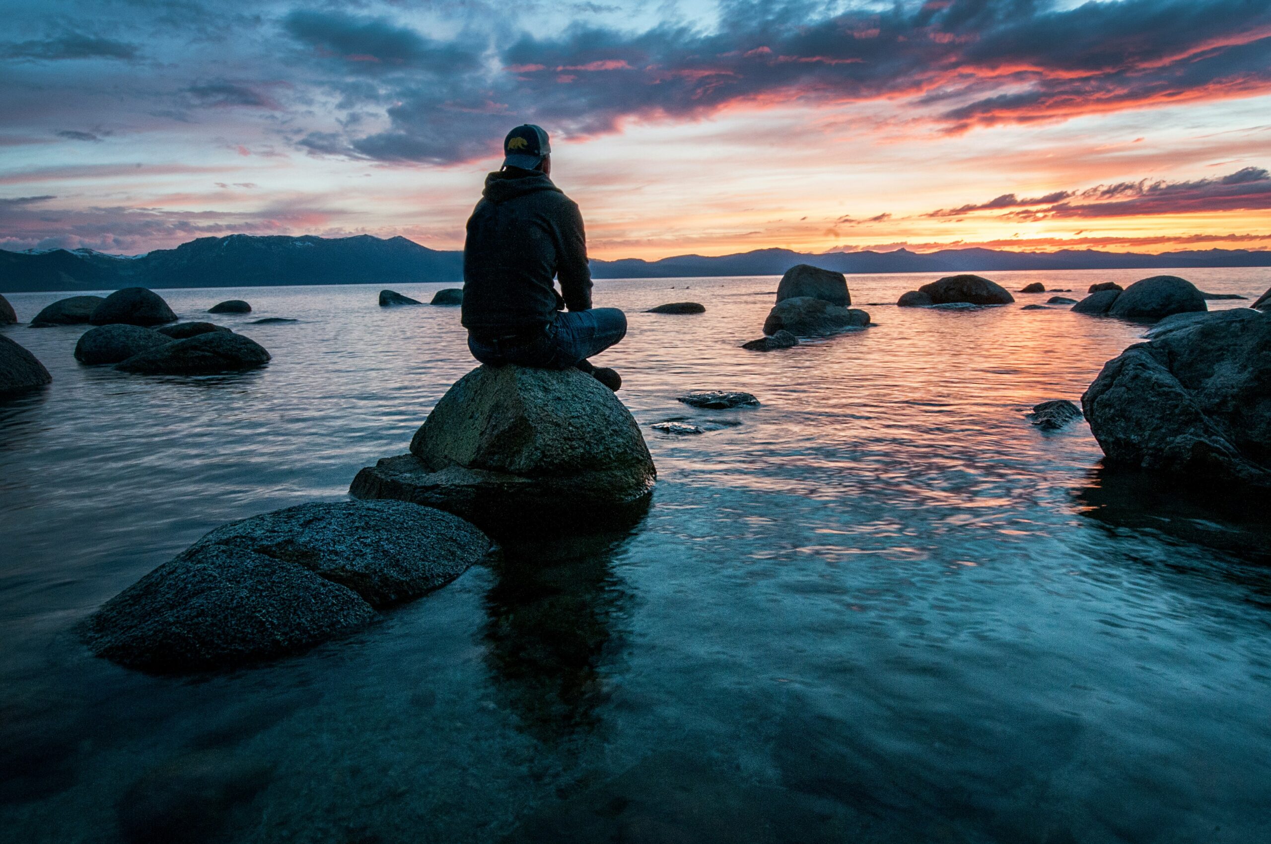 A person sitting on a rock by a calm lake at sunset, reflecting in a peaceful setting.