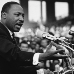 Black and white image of Martin Luther King Jr. passionately delivering a speech at a public event, with microphones in front of him and a crowd in the background.