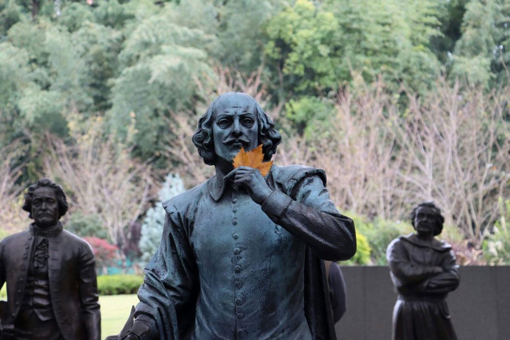 A bronze statue of William Shakespeare holding a leaf, surrounded by trees and other historical figures.