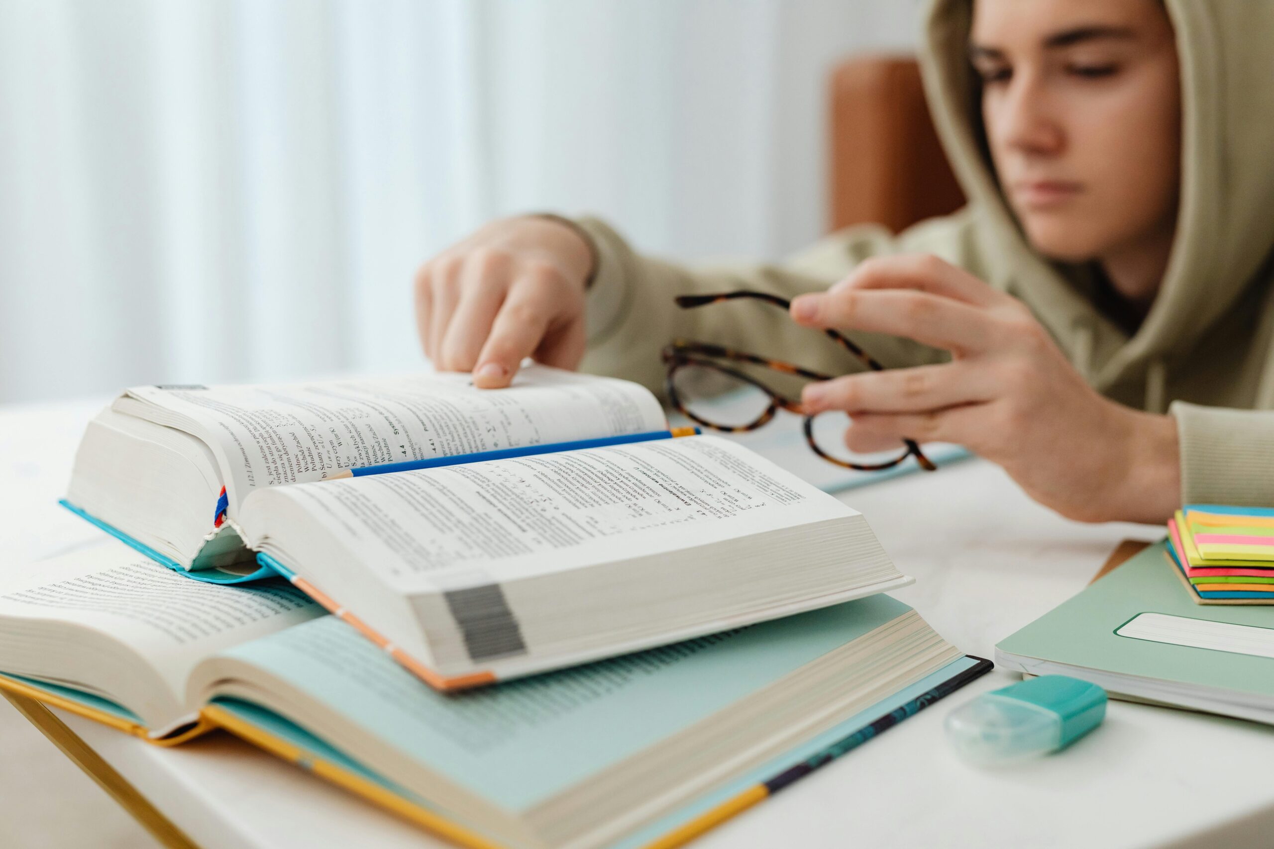 A student in a hoodie studying at a desk with open textbooks, sticky notes, and stationery.