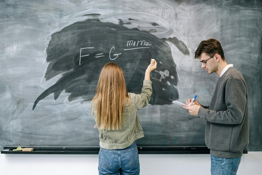 Student writing a formula on a chalkboard while another takes notes.