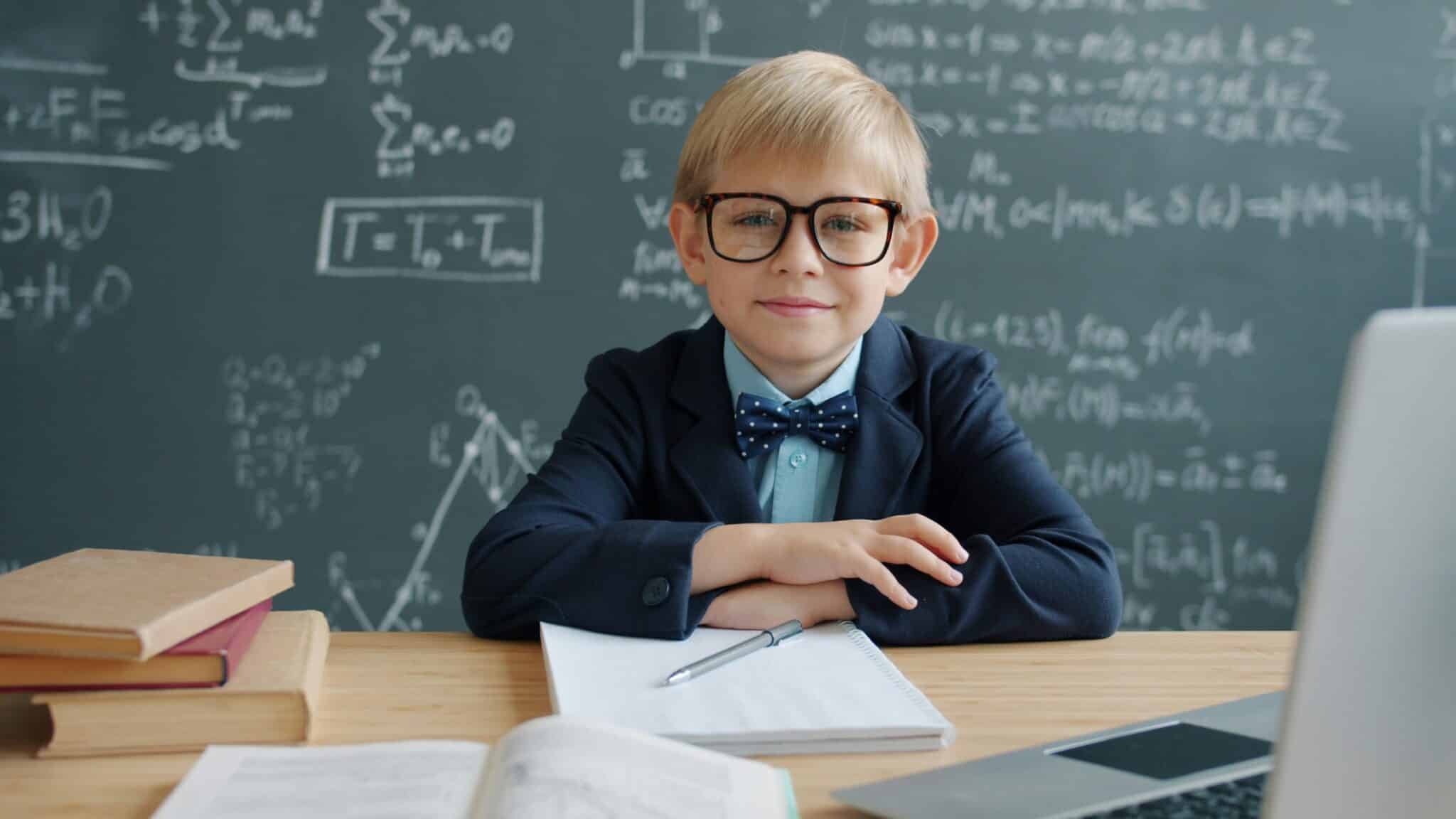 Young student sitting at a desk with textbooks and a laptop in front of a blackboard covered in mathematical formulas and equations