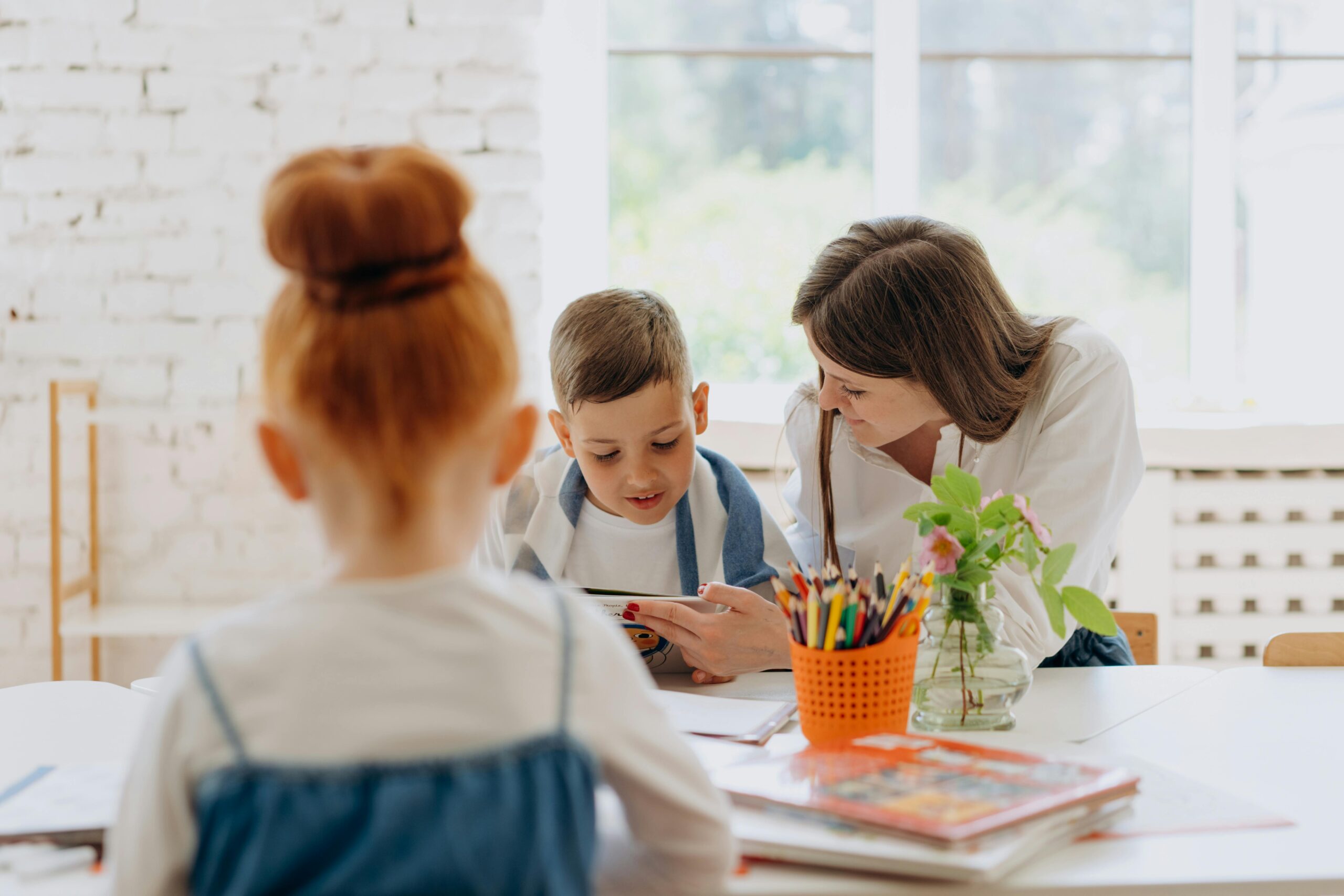 A teacher helps a child with a learning activity while other children are engaged in the background. The teacher and child are focused on a book, surrounded by school supplies.
